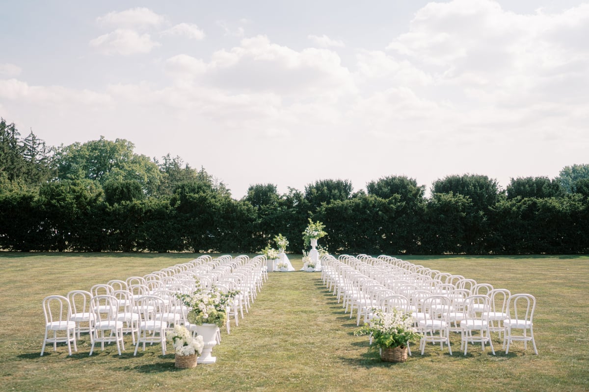 Wide outdoor ceremony aisle lined with elegant wooden folding chairs leading to a floral arch on an expansive lawn framed by trimmed greenery