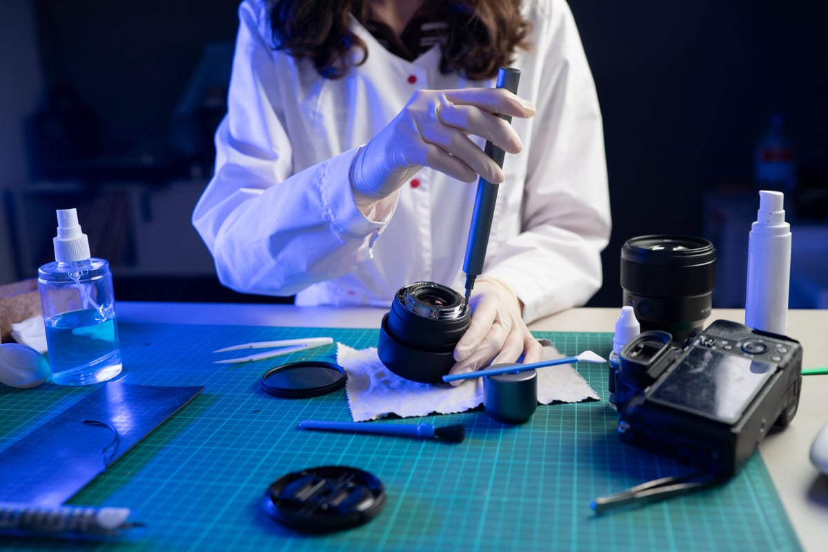 A person repairs a camera lens using tools and cleaning supplies on a workshop table at night