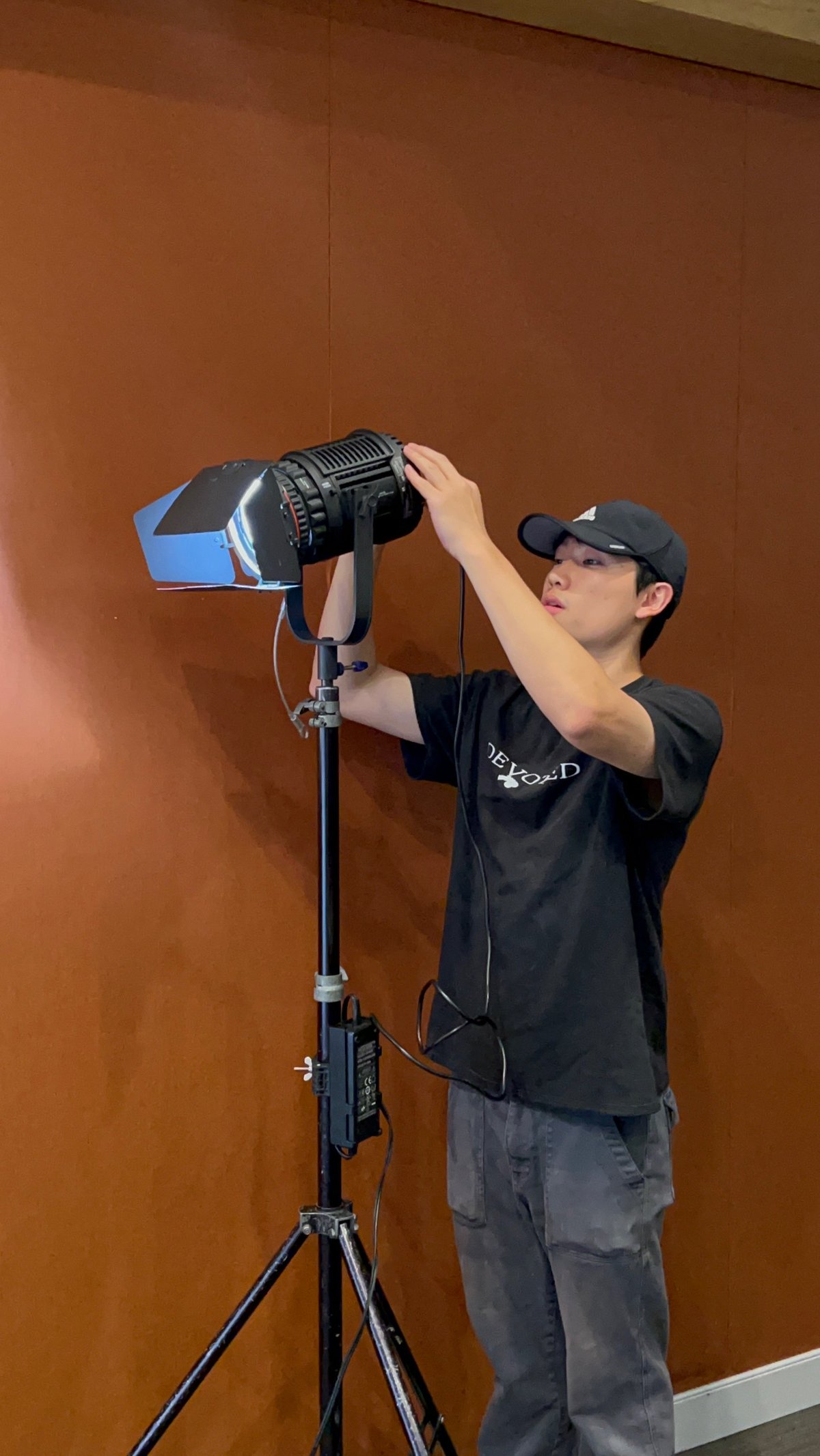 Crew member in black DEVOTED shirt and cap adjusting a professional film light on a tripod against an orange wall