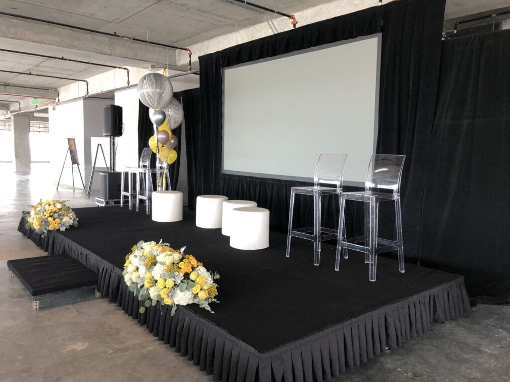 Black stage setup with projection screen, yellow and white floral arrangements, bar seating, and draped black platform for event or presentation