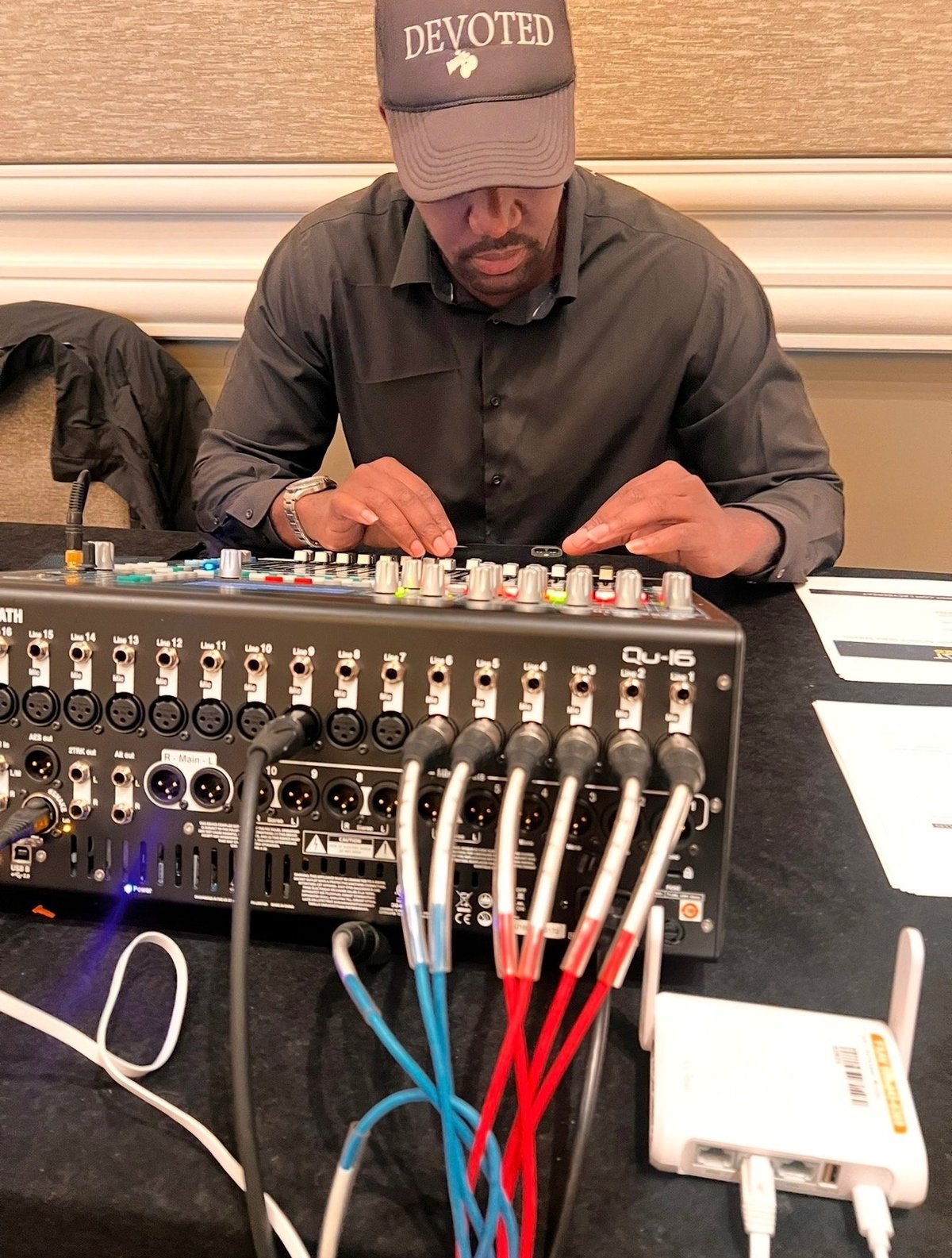 Crew member in a DEVOTED cap operating an audio mixing console with multi-colored XLR cables