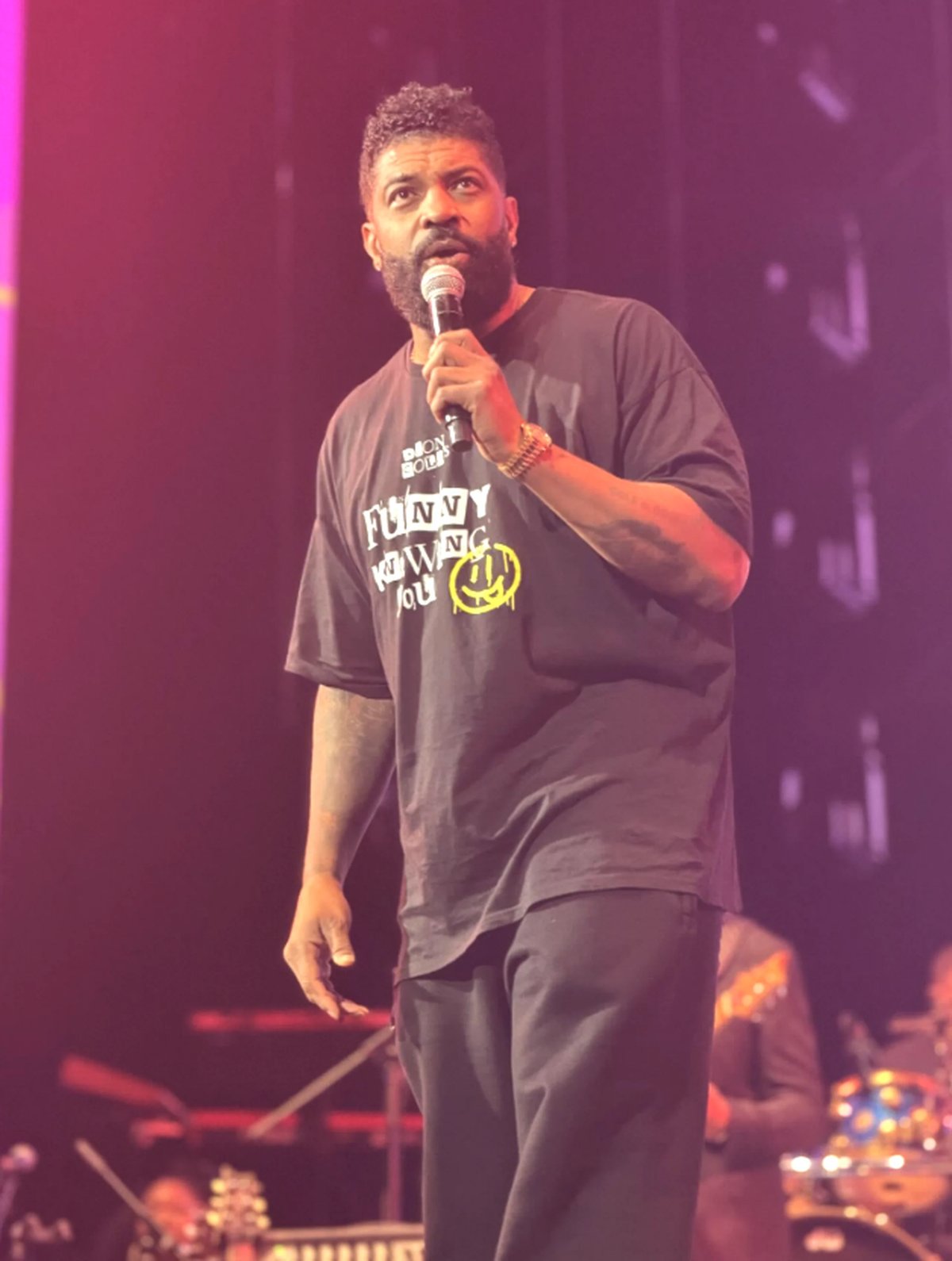 Man performing on stage holding a microphone against a purple-lit background, wearing a dark graphic t-shirt