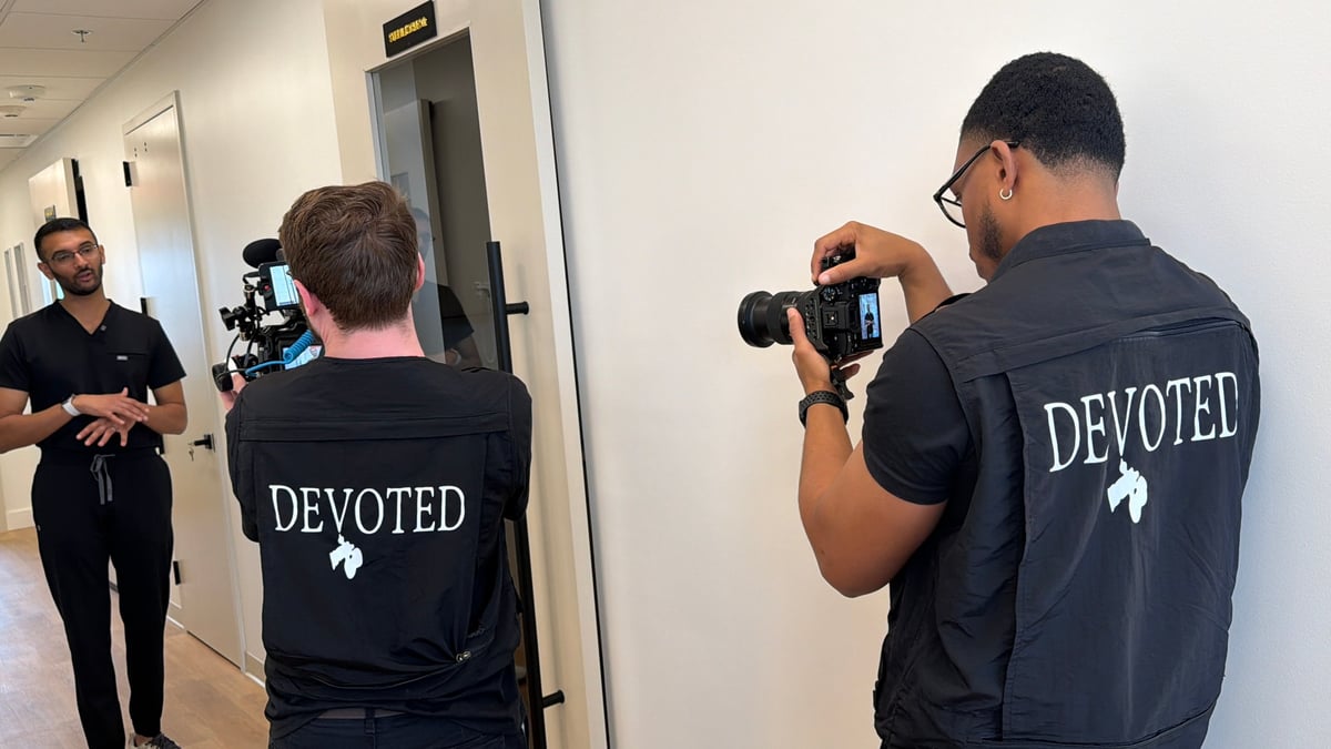Two people wearing black DEVOTED shirts recording video indoors, with a man in black shirt standing in the hallway to the left