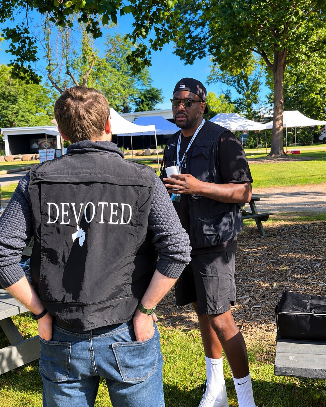 Man wearing DEVOTED vest converses with another man outdoors in a park setting during daytime
