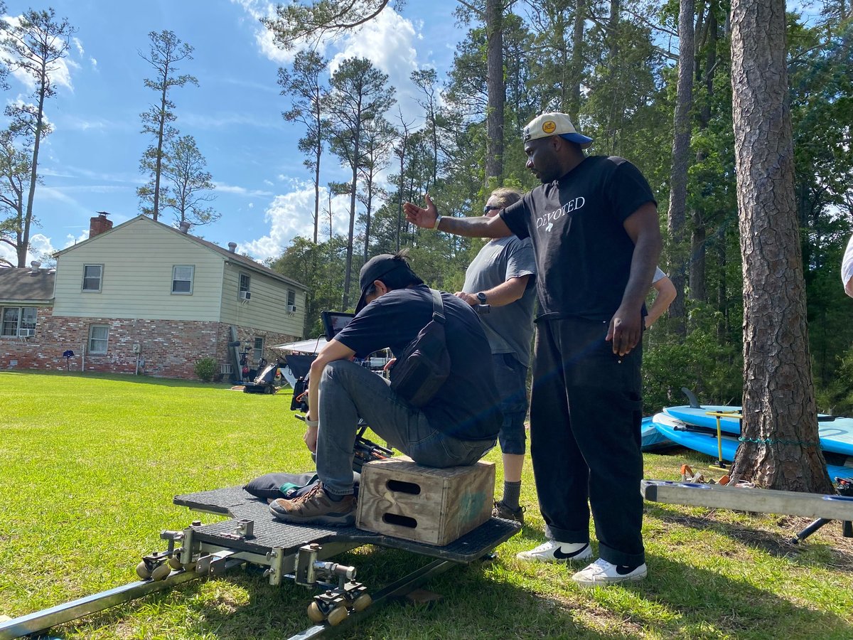 Group of crew members working on camera equipment in a grassy yard with trees and a house in the background