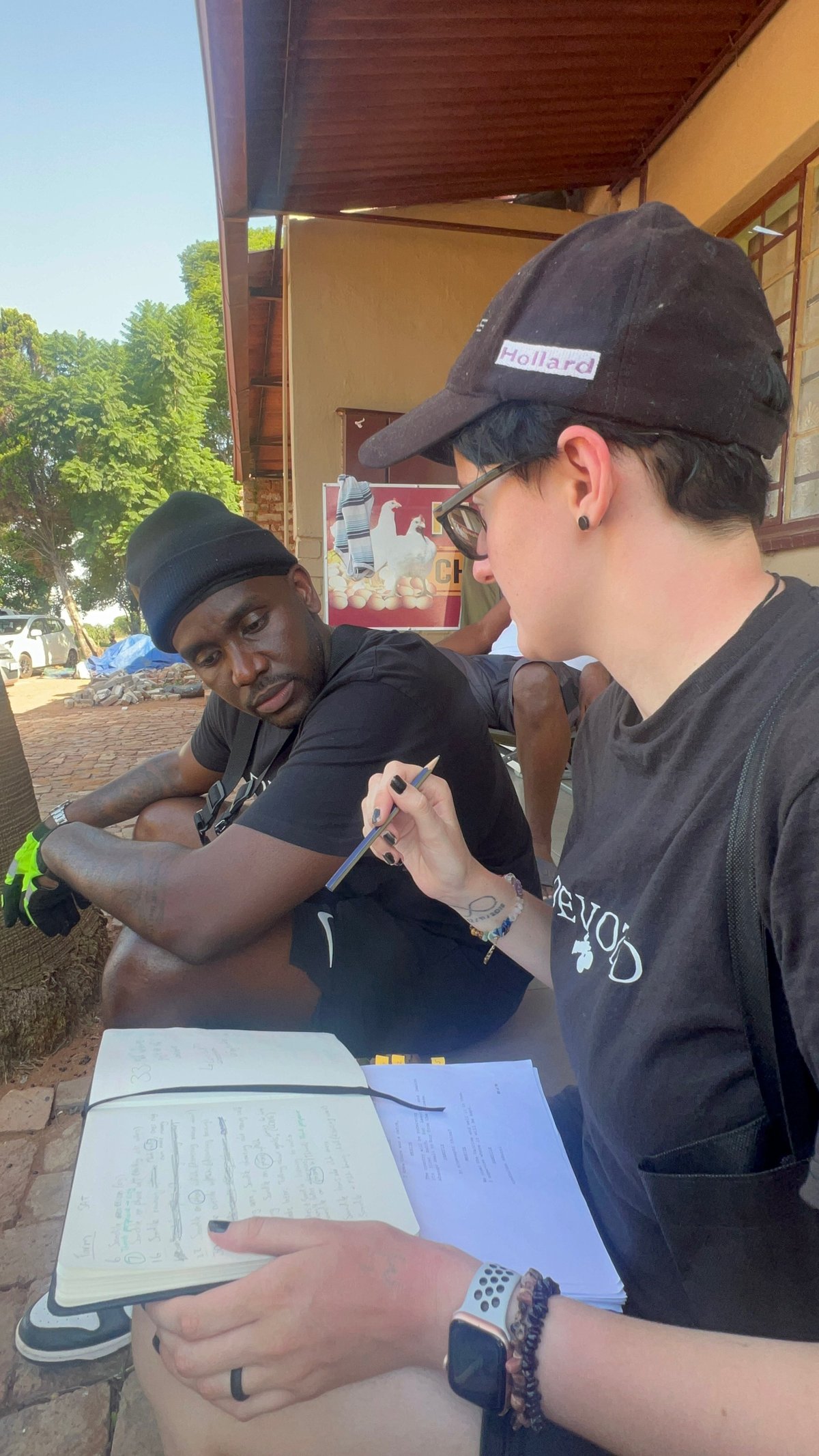 Two crew members reviewing documents and shot plans at an outdoor table in front of a wooden structure, with green trees in the background