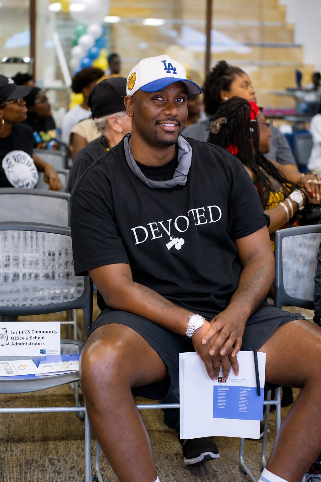 Man wearing LA cap and Devoted shirt sits on chair holding a program at an indoor community event