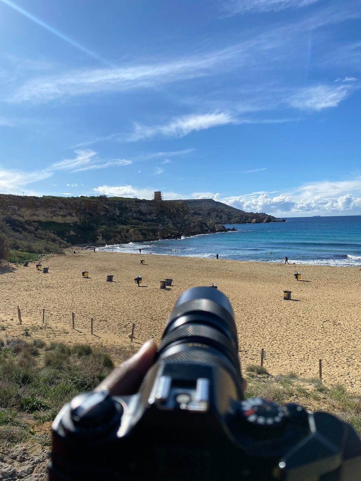 Person holding a camera pointed at a scenic beach cove with turquoise water, sandy shore, and distant cliffs under blue sky