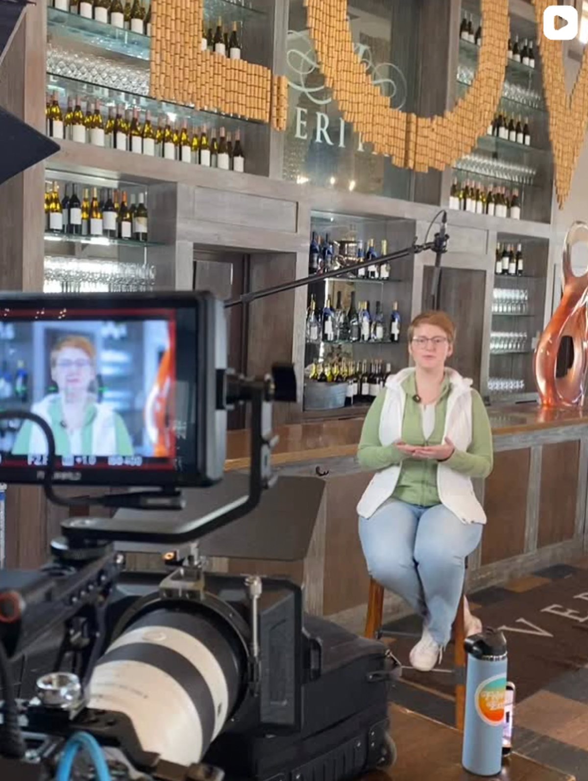 Woman speaking on camera in wine bar with brick wall and bottles in background