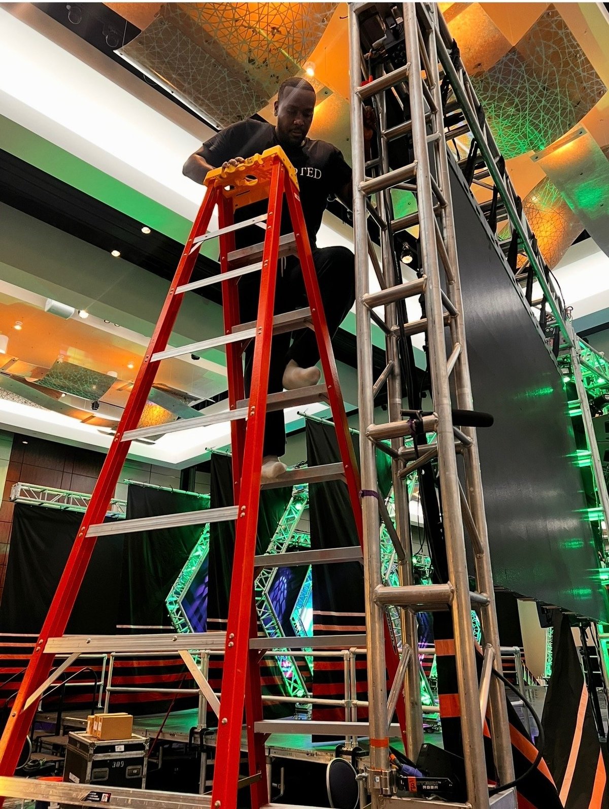 Technician on a red ladder installing stage lighting under orange and green overhead panels