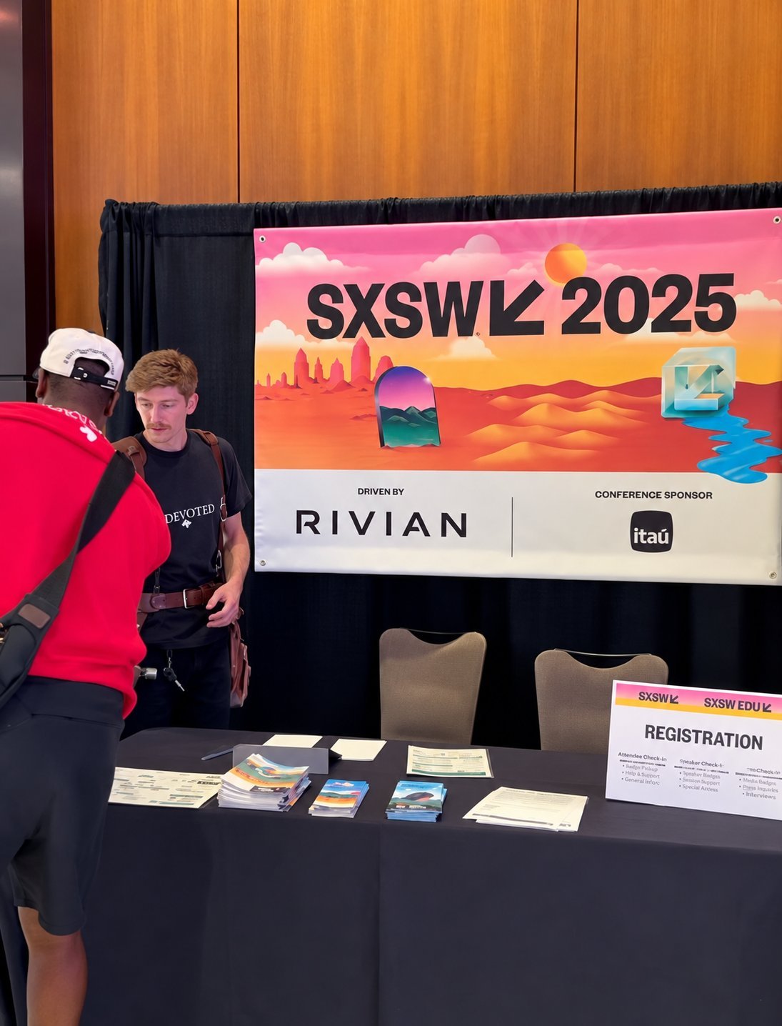 SXSW 2025 conference registration area with branded banner on the wall, registration table with materials, and two attendees, one wearing a black DEVOTED shirt