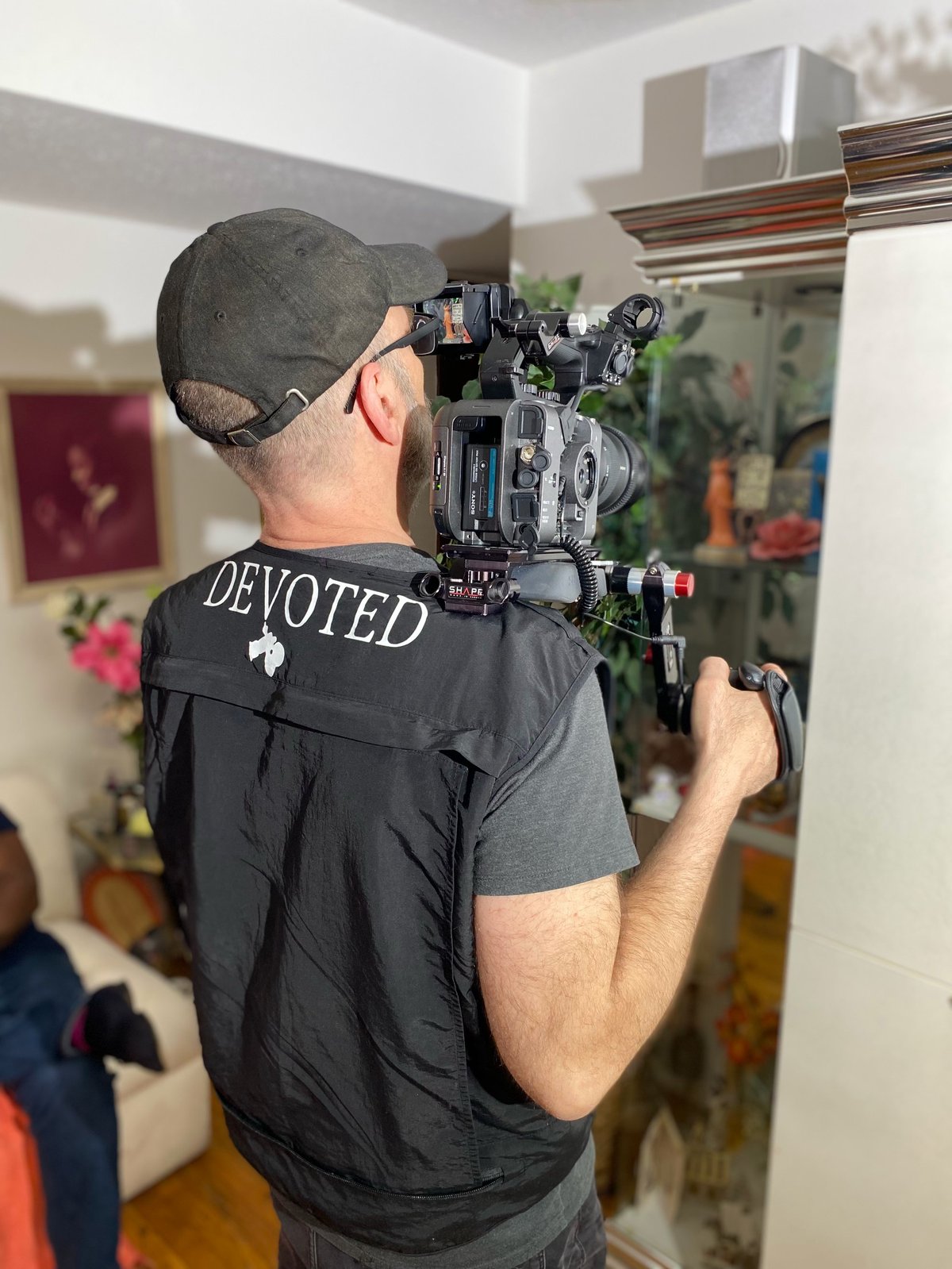 Cameraman wearing a black DEVOTED vest and cap, operating a professional cinema camera rig in an indoor room with plants and mirror