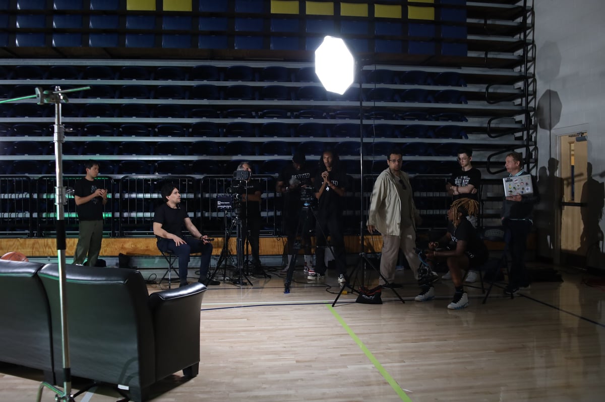 Film production crew gathered under bright octagonal lighting on a wooden gymnasium stage with empty bleachers behind them