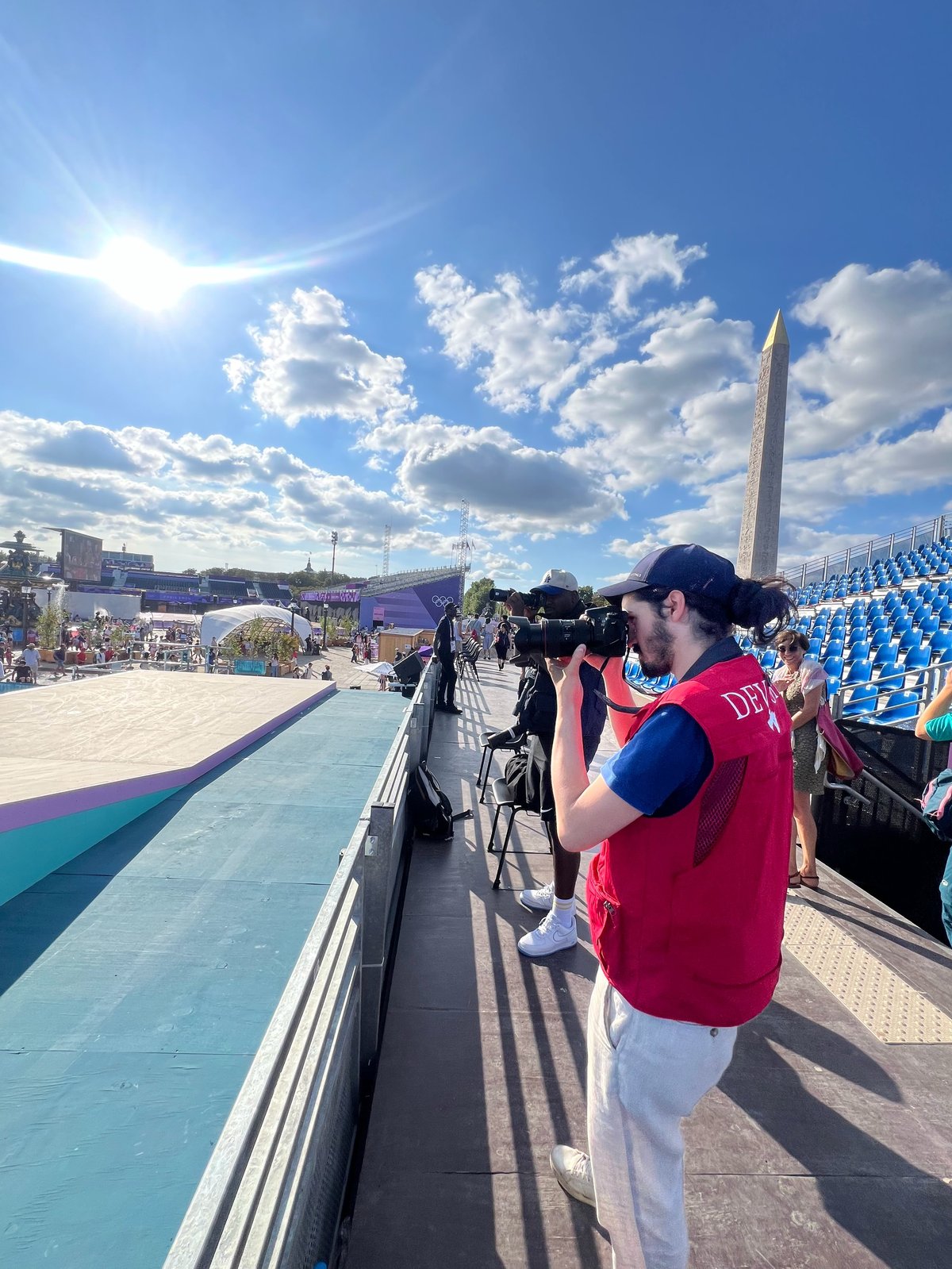 Photographer in red DEVOTED vest and white pants shooting from bleachers overlooking the Paris 2024 venue with the Washington Monument visible in background