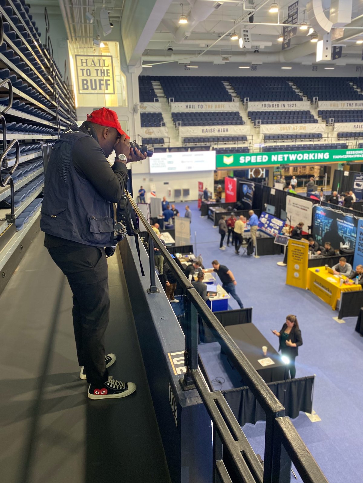 Person in red hat and black DEVOTED jacket leaning over arena railing, photographing event floor with vendor booths and attendees below