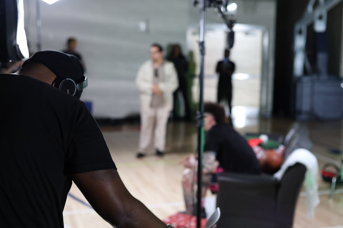 Crew member in black shirt wearing headphones with arm raised, operating camera equipment in professional studio setting with cast and lighting in background
