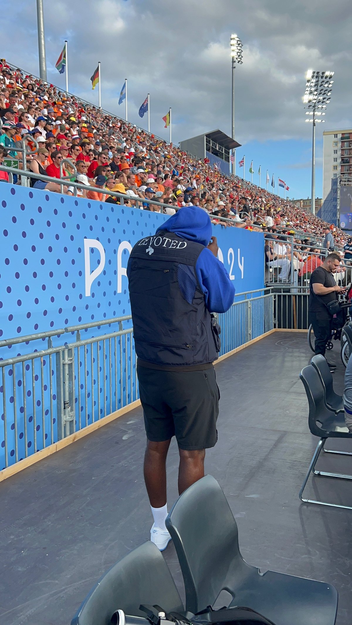 DEVOTED crew member in navy jacket on stadium track with packed stands and international flags in the background