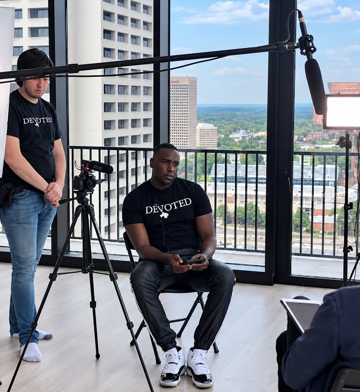 Two people conducting a video interview on a high-rise balcony overlooking a cityscape with mountains in the distance