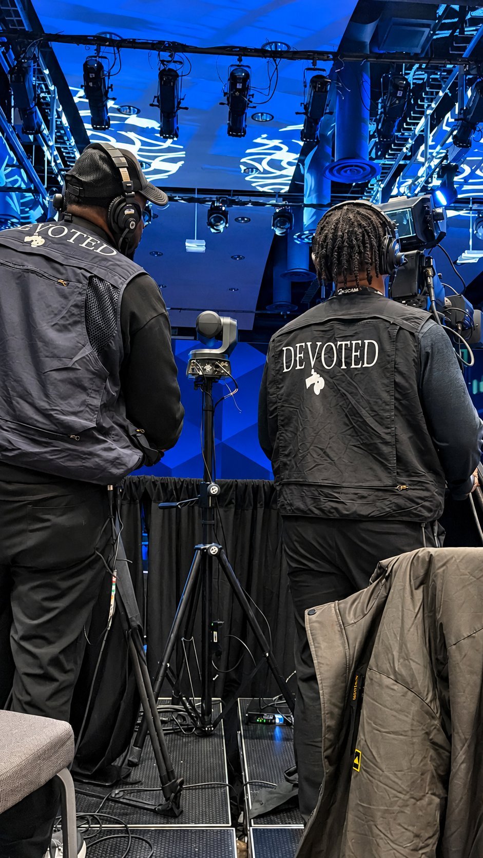 Two crew members wearing DEVOTED jackets operate camera equipment on a film set with blue lighting and overhead rigs