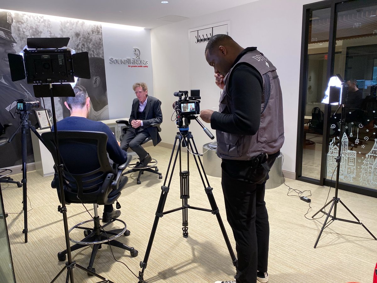 Professional video production setup in studio with cameraman operating equipment, interviewer seated, and lighting rigs positioned around the set