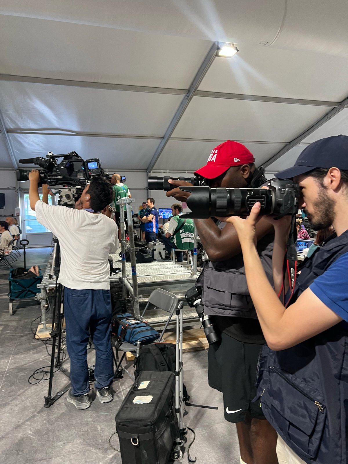 Film crew operating broadcast cameras under a tent at an outdoor event, with three cameramen recording footage and equipment visible