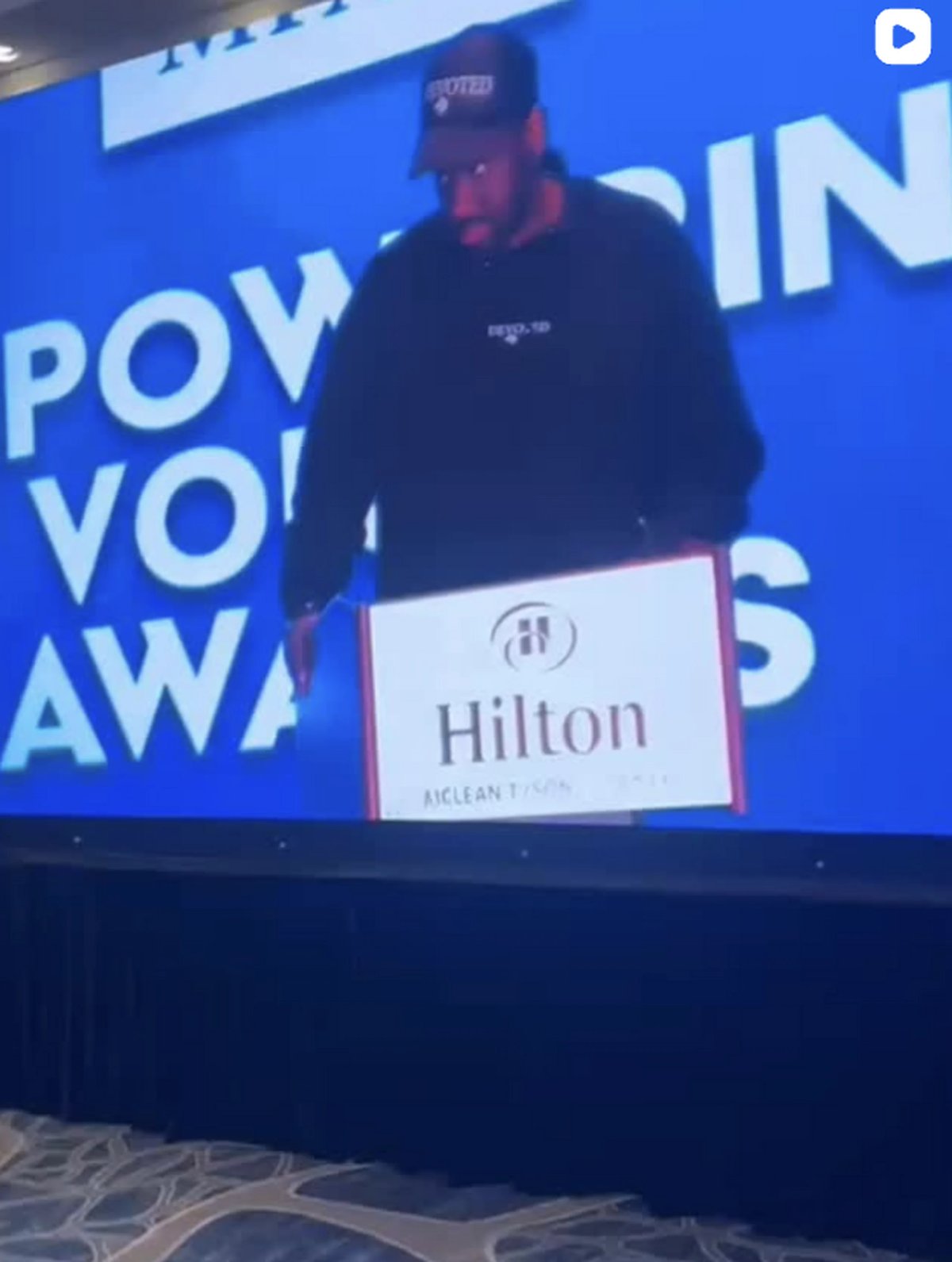 Person speaking at awards show podium with Hilton branding on blue background with partial text visible