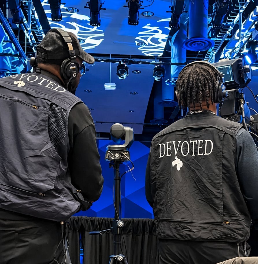 Two crew members wearing black DEVOTED jackets stand beneath professional camera and lighting rigs in a blue-lit studio