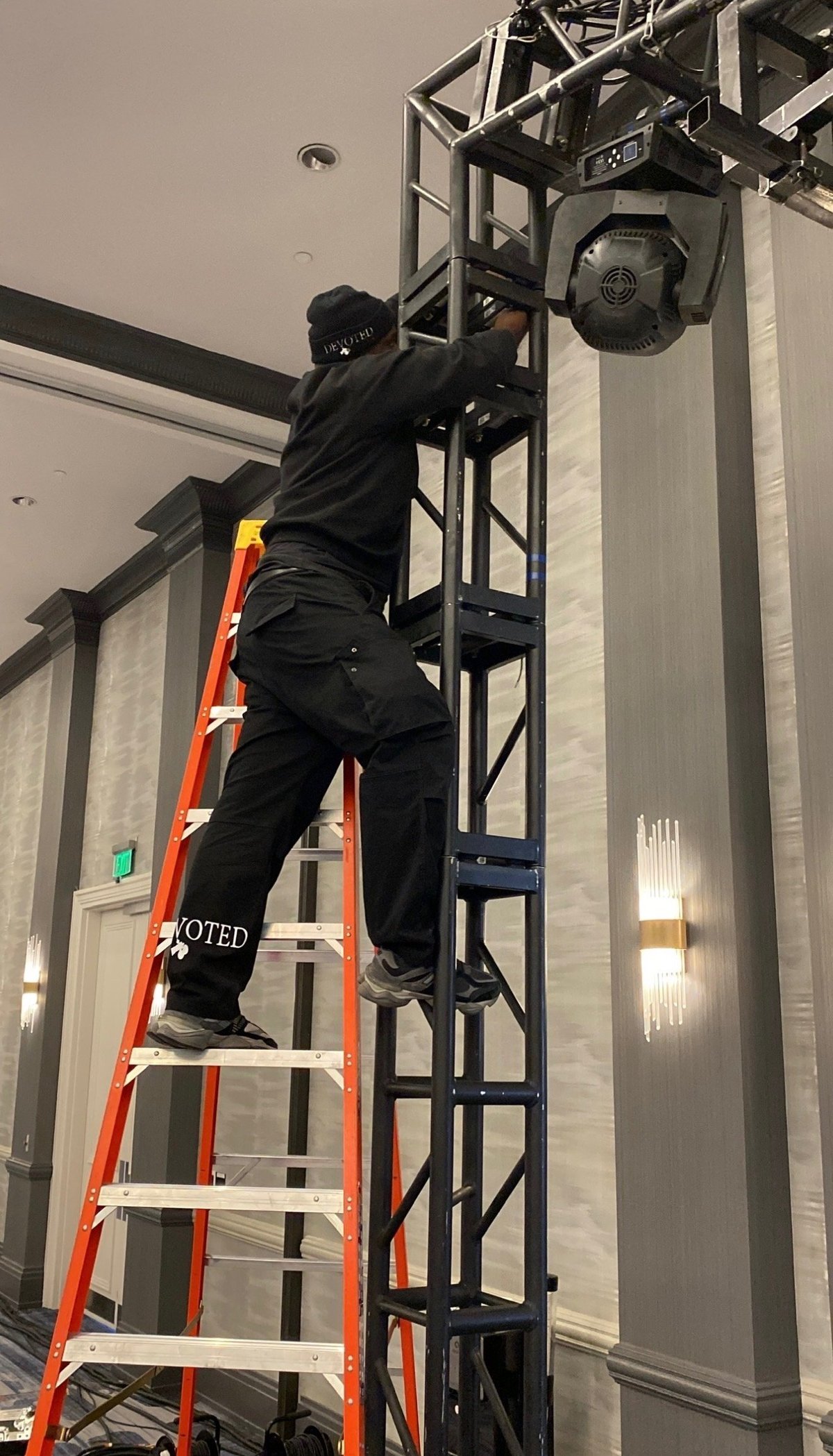 Crew member in a DEVOTED beanie climbing an orange A-frame ladder to rig lighting on a truss grid