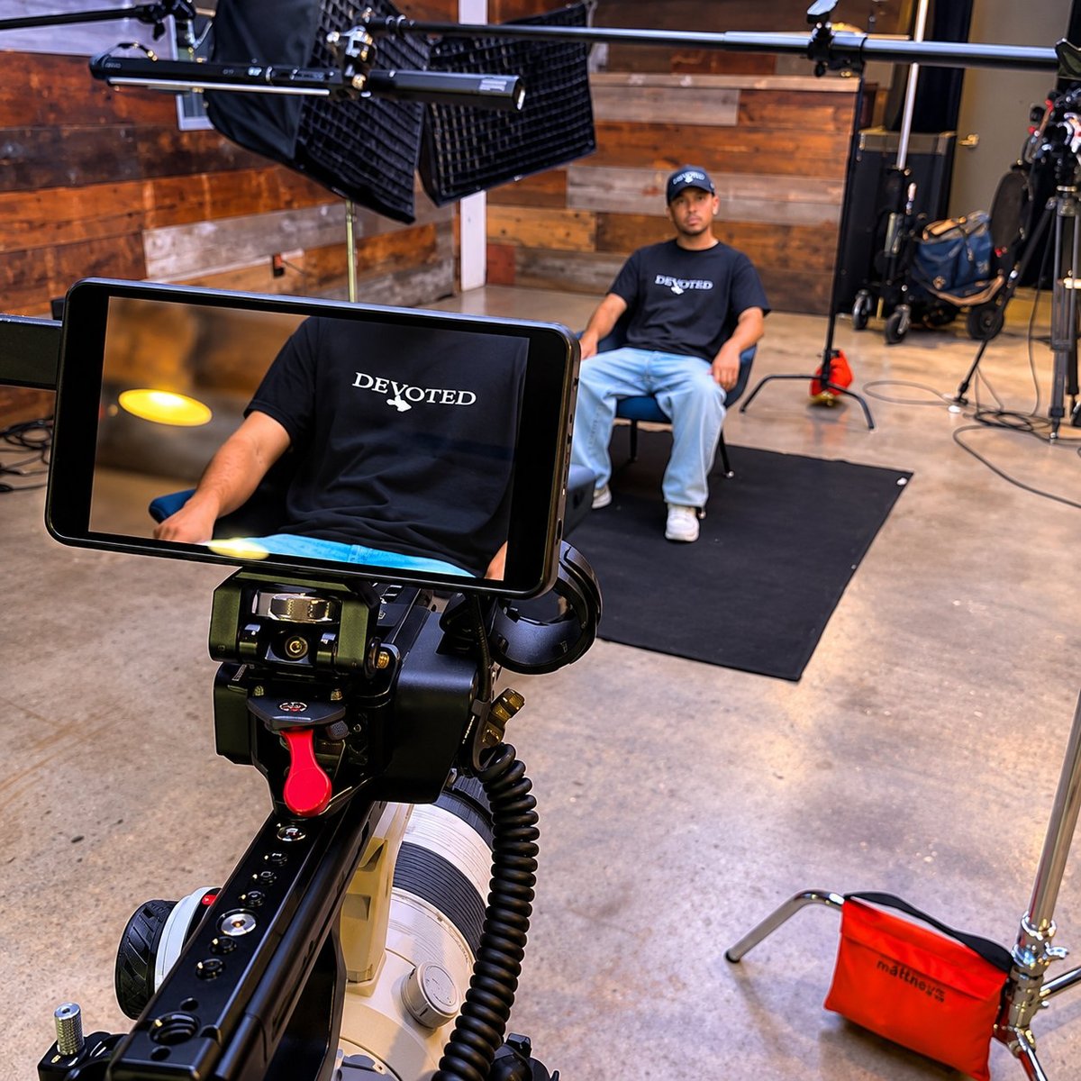 Film production set with a cinema camera on a tripod showing a subject in a DEVOTED shirt on its field monitor, with a seated subject under professional lighting gear in the background