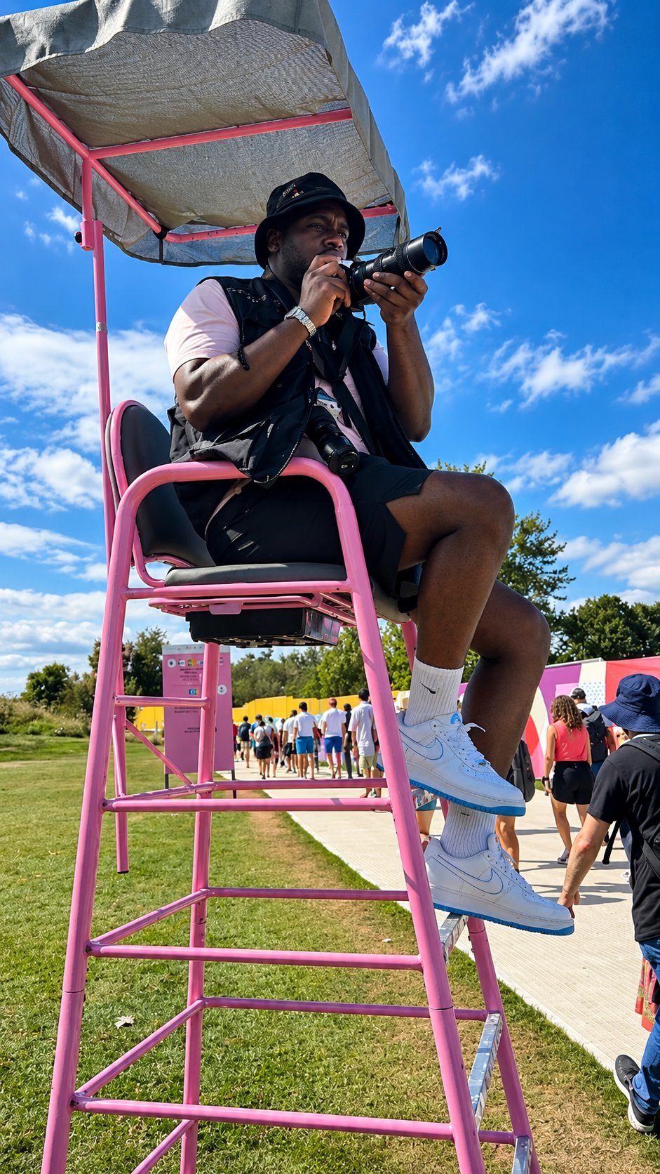 Person sitting on a tall pink lifeguard stand with a camera, overlooking an outdoor event with a crowd