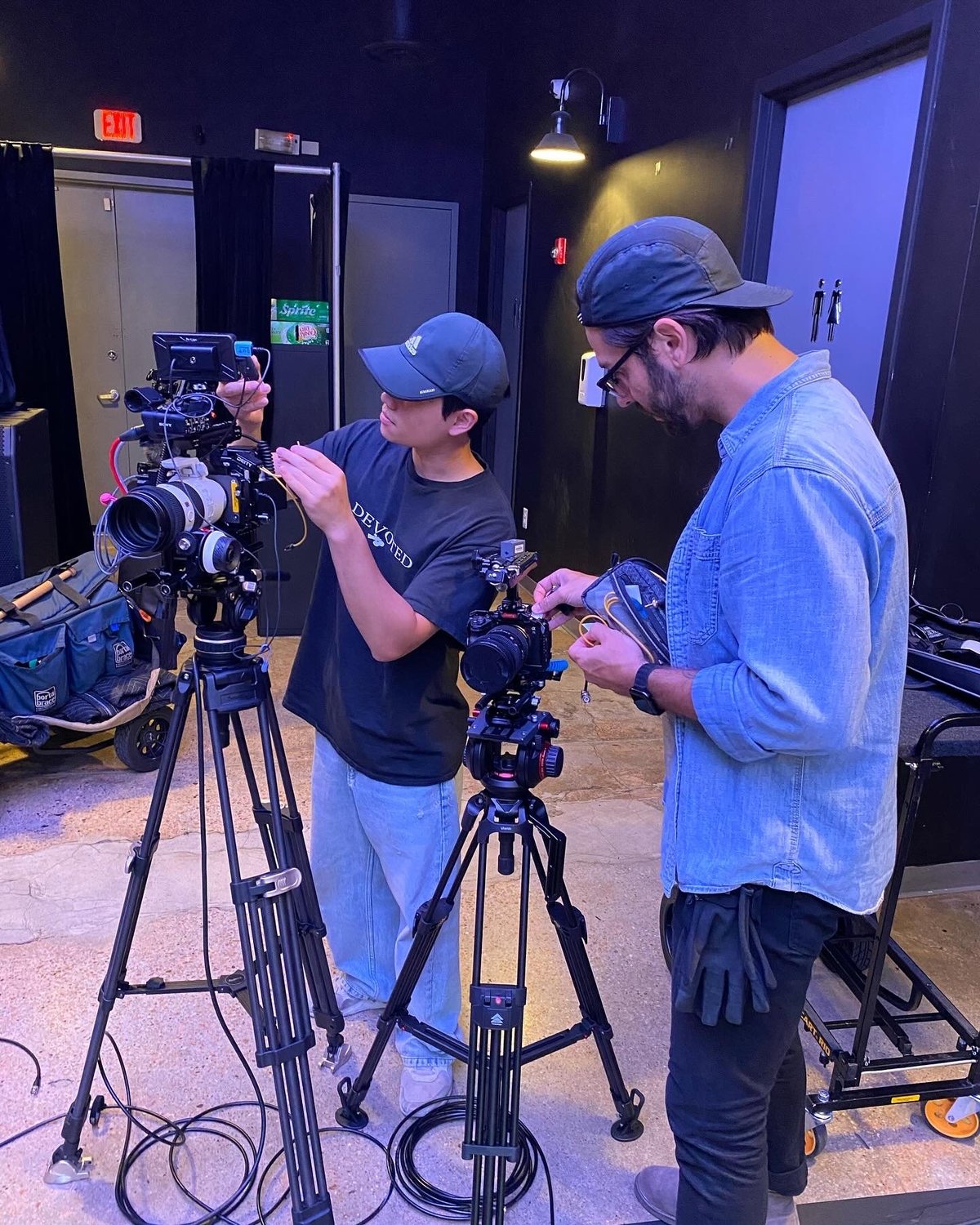 Two camera operators in blue caps adjusting professional video equipment on tripods in a dark studio setting