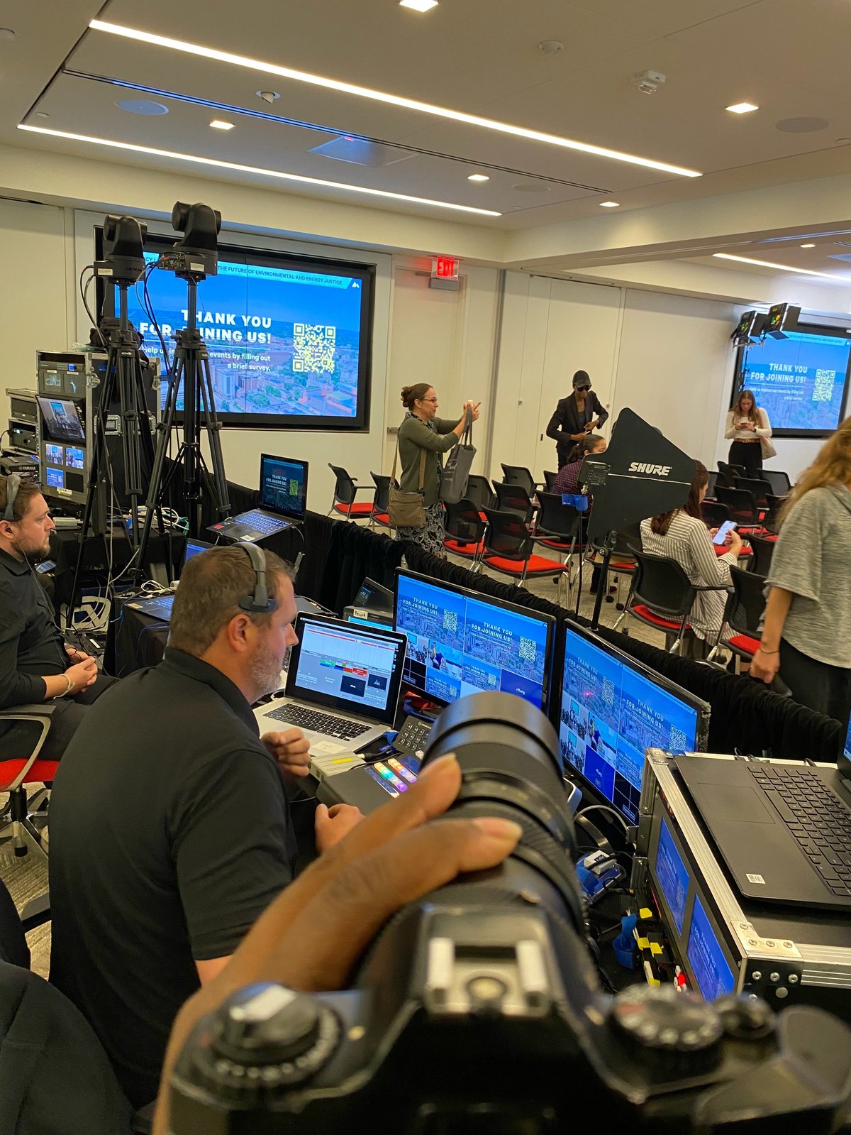 Production crew filming a live event in a studio with cameras, monitors, and technical equipment, people visible on stage and at work stations